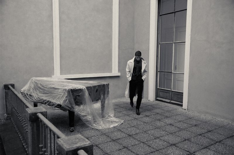 Man wearing a light overcoat and dark trousers walks across a stone courtyard beside a grand piano covered in transparent plastic film.