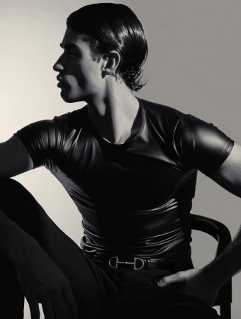Niccolo Fortini in profile wearing a tight black short-sleeve top and belt with a silver horsebit buckle while seated in studio.