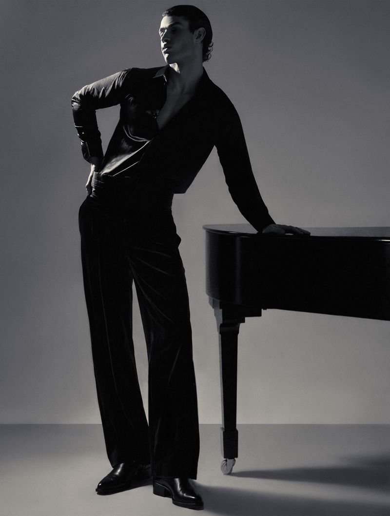 A man in an unbuttoned black shirt and wide-leg trousers standing and leaning against a black grand piano in a studio.