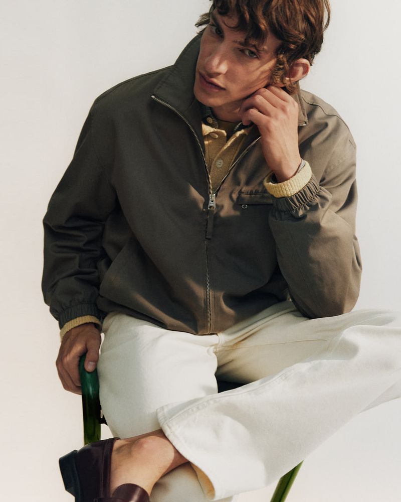 Seated man wearing a taupe zip-up jacket, cream knit shirt, and white jeans on a green chair against a white background.