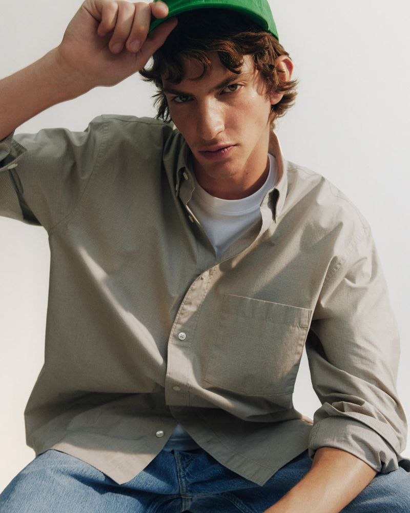 Close-up portrait of a male model wearing a green cap, beige button-down shirt, and blue jeans against a neutral studio backdrop.