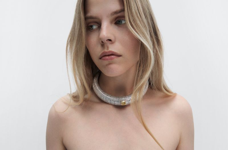 Close-up studio shot of a young blonde woman wearing a silver and gold tone necklace against a plain white backdrop. Minimalist fashion aesthetic.