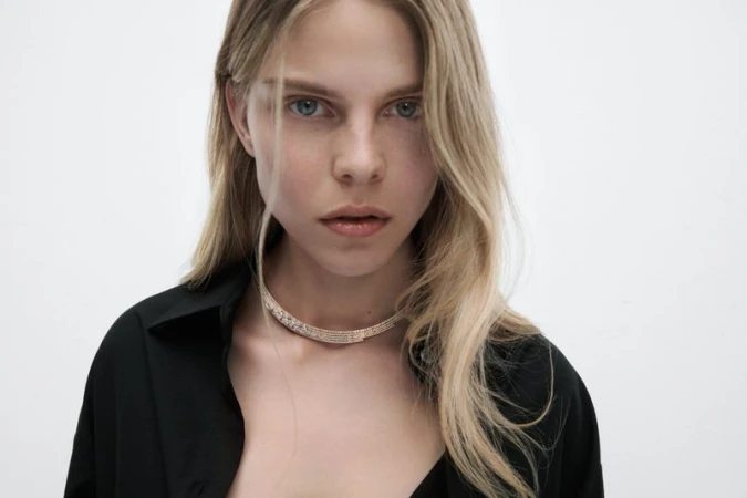 Close-up portrait of a fair-skinned young woman with blonde hair wearing a jeweled choker and black button-down shirt against a white background.