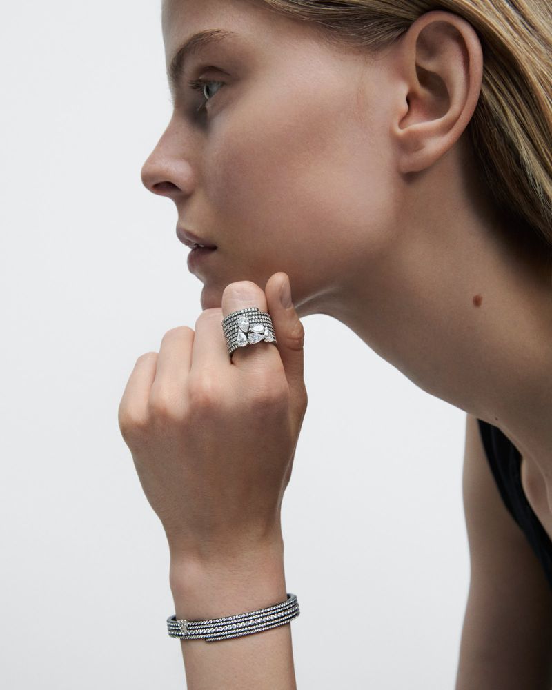 Woman in profile showcases crystal jewelry: a wide band ring with large clear stones and a delicate, matching bracelet, against a plain background.