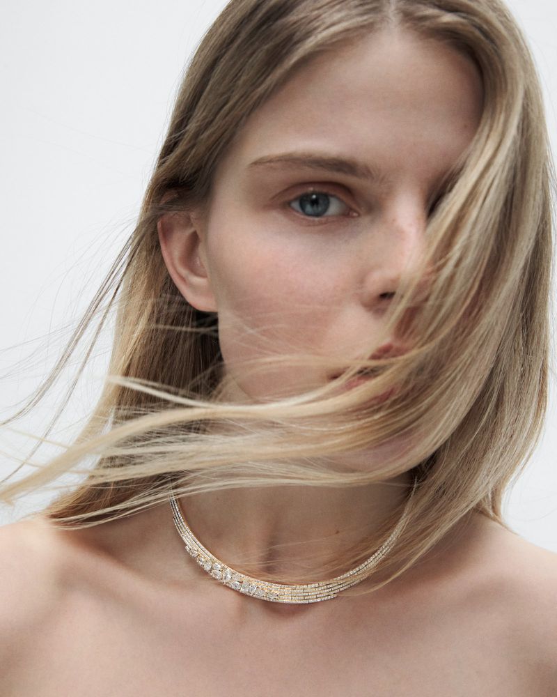 Close-up of an adult woman with fair skin and blonde hair wearing a jeweled necklace against a neutral backdrop in a studio setting.