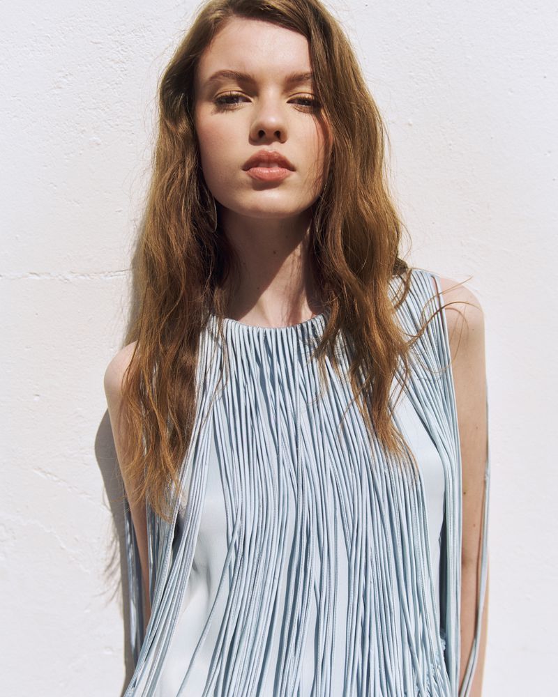 A woman with wavy brown hair posing against a sunlit white textured wall wearing a light blue pleated fringe sleeveless top.
