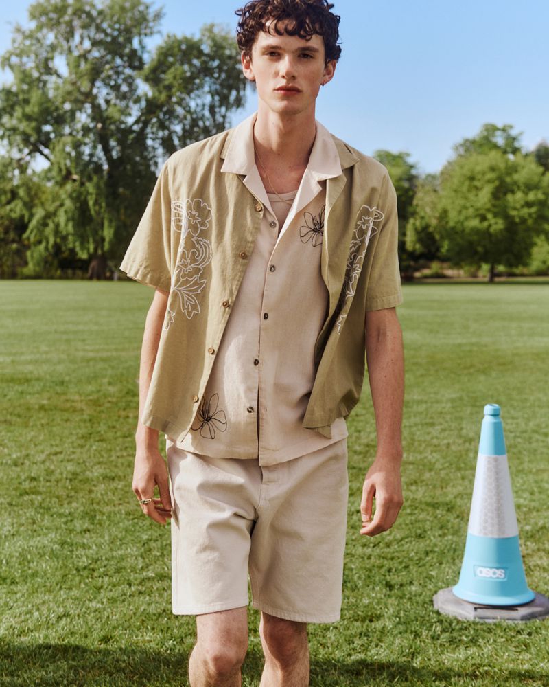 Young man standing on a grassy park lawn wearing an embroidered olive camp shirt and beige shorts under bright natural daylight.
