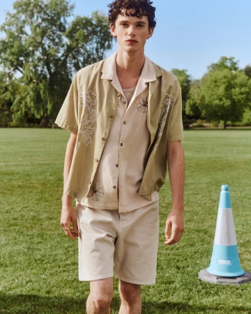 Young man standing on a grassy park lawn wearing an embroidered olive camp shirt and beige shorts under bright natural daylight.
