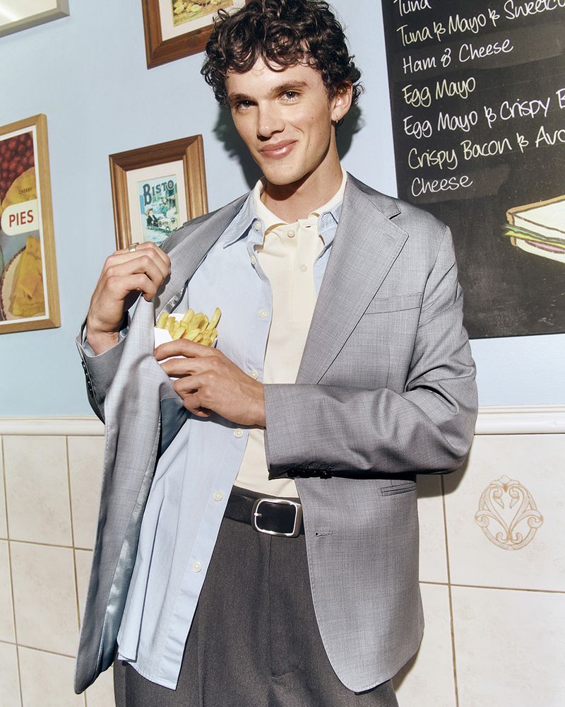 Young man wearing a grey blazer, blue shirt, and holding fries inside his jacket pocket in a cafe.