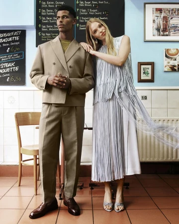 A man in a beige double-breasted suit and a woman in a pale blue fringe dress posing together inside a cafe.