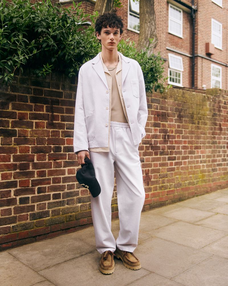 A young male model posing outdoors wearing a white jacket, trousers, and beige shoes while holding a black cap.