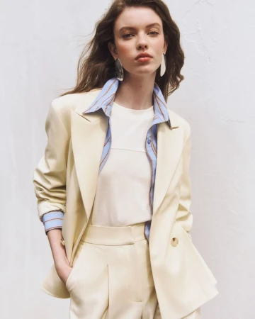 Model wearing a pale cream tailored suit, striped blue shirt, white t-shirt, and silver earrings against a neutral backdrop.