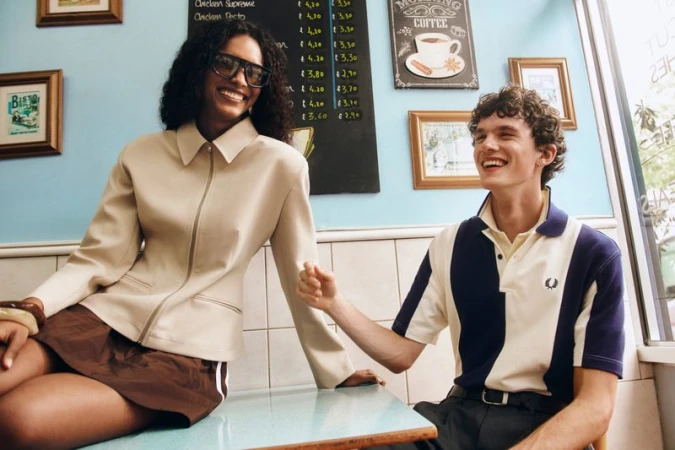 A woman in a cream zip jacket and dark glasses sits on a cafe table beside a man in a navy and cream polo shirt laughing.