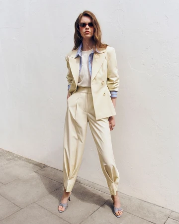 Model wearing a pale yellow tailored suit with sunglasses and open-toe heels, standing against a white wall outdoors.