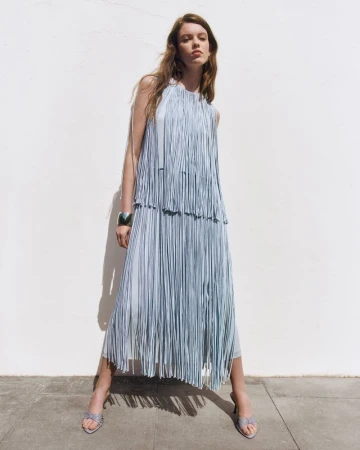 Model wearing a light blue fringed sleeveless dress, silver cuff bracelet, and heeled sandals against a white outdoor wall.