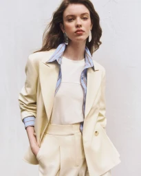Model wearing a pale cream tailored suit, striped blue shirt, white t-shirt, and silver earrings against a neutral backdrop.