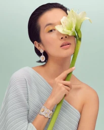 Model Kate Tsai holding a white lily stem, wearing a pleated one-shoulder top and floral silver jewelry against a pale mint background.