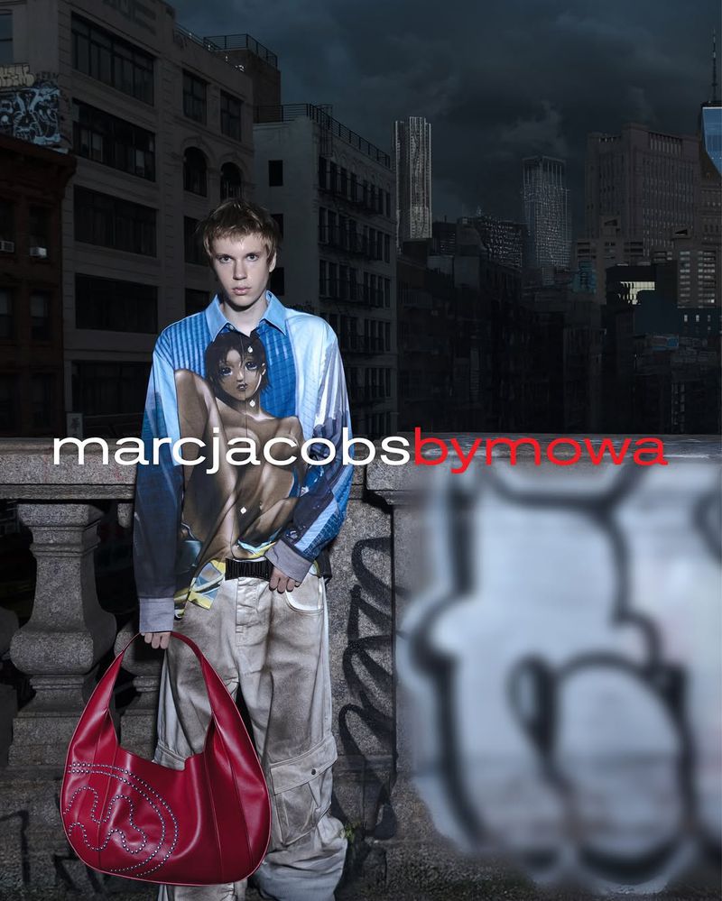Young adult man in a patterned shirt and cargo pants stands on a city terrace holding a red handbag, buildings in the background.