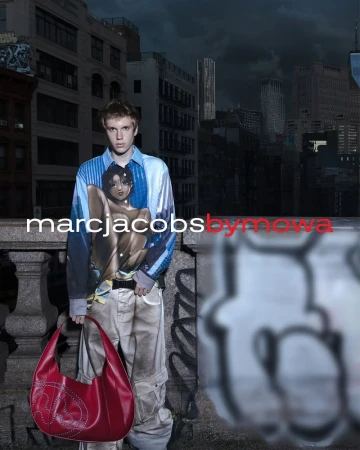 Young adult man in a patterned shirt and cargo pants stands on a city terrace holding a red handbag, buildings in the background.