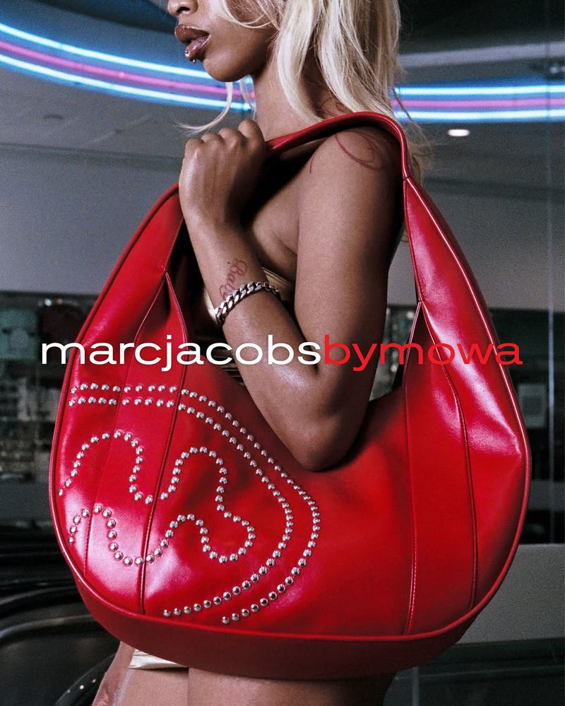 A woman poses with a red, studded Marc Jacobs by Mowa handbag, featuring visible branding against a soft background with neon lighting.