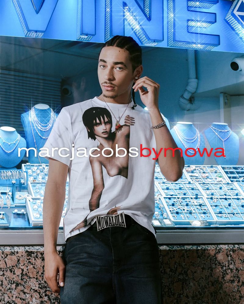 Male fashion model poses in front of a jewelry store window wearing a white graphic t-shirt, jeans, and jewelry in a studio setting.