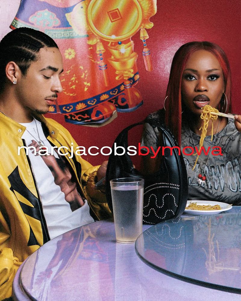 Two people sit at a table eating noodles, one using chopsticks. The table is round and lilac-colored. Logos for Marc Jacobs visible.