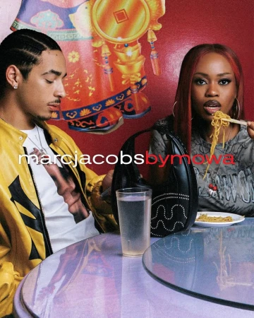 Two people sit at a table eating noodles, one using chopsticks. The table is round and lilac-colored. Logos for Marc Jacobs visible.