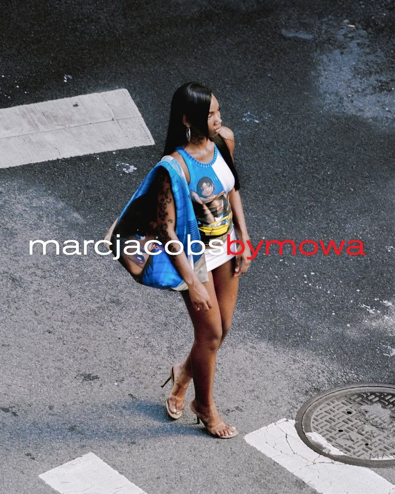A slender dark-skinned woman strides across a crosswalk in the city, wearing high heels and carrying a blue Marc Jacobs tote bag.