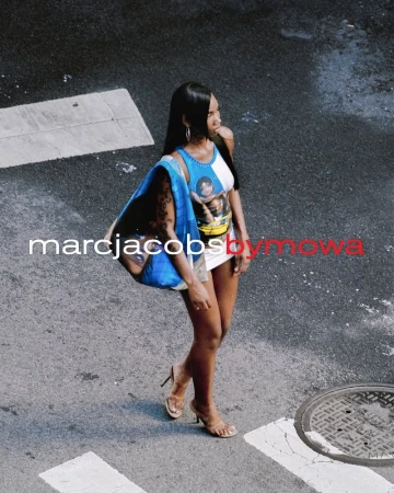 A slender dark-skinned woman strides across a crosswalk in the city, wearing high heels and carrying a blue Marc Jacobs tote bag.
