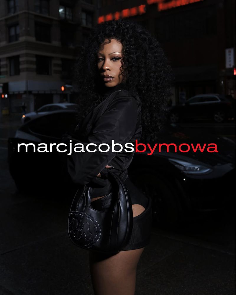 Woman in a black outfit poses with a purse on a city street at night, Marc Jacobs by Mowa logo visible.