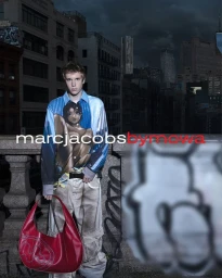 Model wearing an anime-printed shirt and beige cargo trousers holding a red leather handbag on an urban rooftop balustrade.