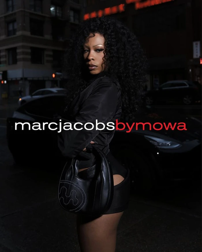 A Black woman with voluminous curly hair posing with a black leather shoulder bag on a city street at night in front of a dark car.