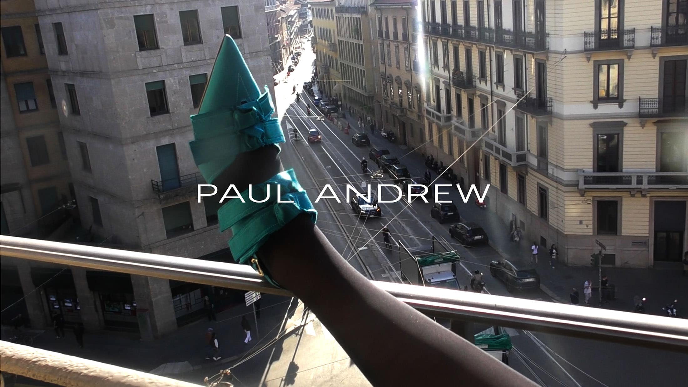 Model's leg in sheer black tights and a bright teal pointed-toe high heel resting on a balcony railing above a city street.