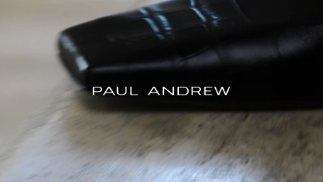 Close-up of a black crocodile-embossed leather square-toe shoe resting on a textured ground with the Paul Andrew logo overlay.