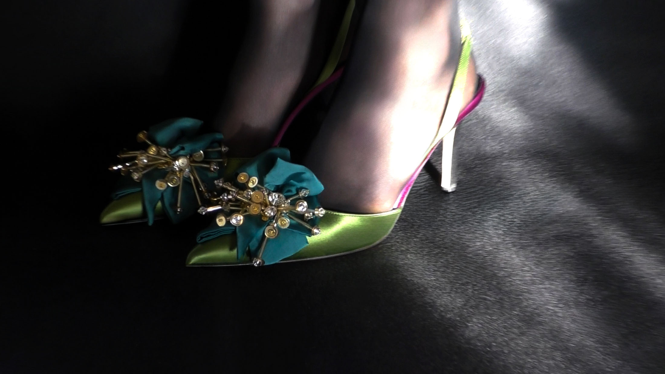 Close-up of feet in sheer black stockings wearing green satin slingback pumps with jeweled teal rosettes under dramatic lighting.