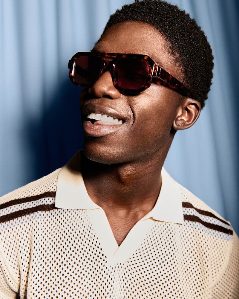 Model Jeremiah Berko Fourdjour smiling in oversized tortoiseshell sunglasses and a textured cream polo shirt against a blue background.