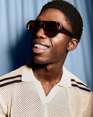 Model Jeremiah Berko Fourdjour smiling in oversized tortoiseshell sunglasses and a textured cream polo shirt against a blue background.