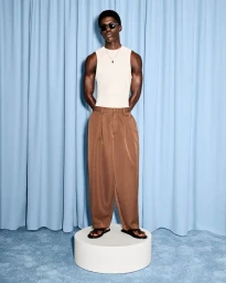 Model Jeremiah Berko Fourdjour in a white tank top, sunglasses, and pleated brown trousers on a white plinth against blue drapery.