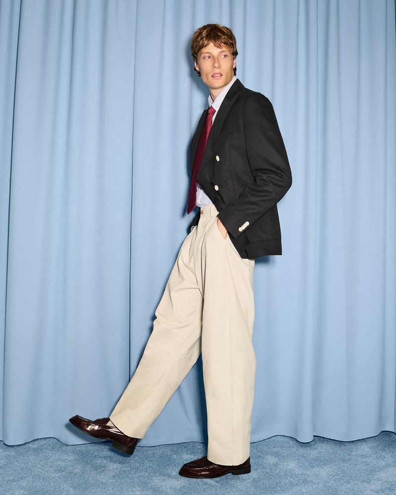 Hugh Laughton-Scott wearing a black blazer, beige wide-leg trousers, and a burgundy tie against a light blue curtain backdrop.