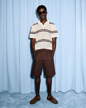 Jeremiah Berko Fourdjour wearing a striped cream knit polo, brown shorts, and sunglasses in front of a blue curtain backdrop.