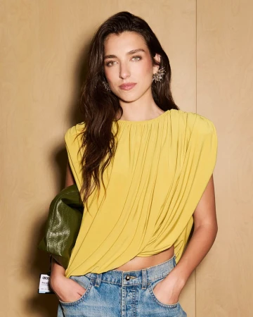 Model Rina Lipa posing in a draped yellow top and blue denim jeans against a light wood panel wall.