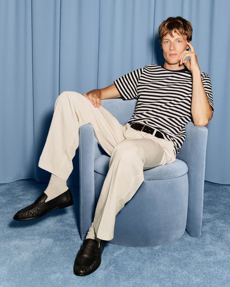 Model Hugh Laughton-Scott in a striped tee and cream trousers seated on a blue velvet armchair against blue draped curtains.