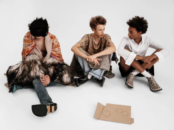 Three male models in contemporary designer menswear seated on a white studio floor beside a flat piece of cardboard.