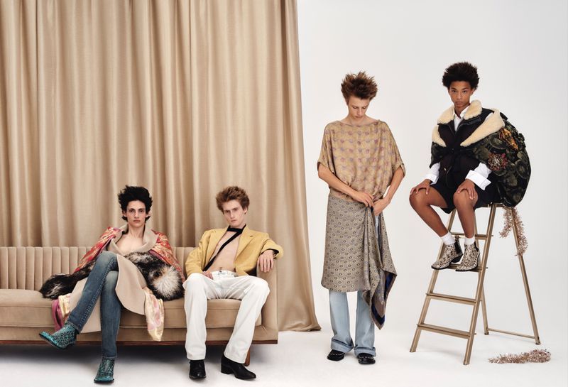 Four male models pose across a tufted sofa and step ladder in eclectic designer menswear against a draped curtain backdrop.