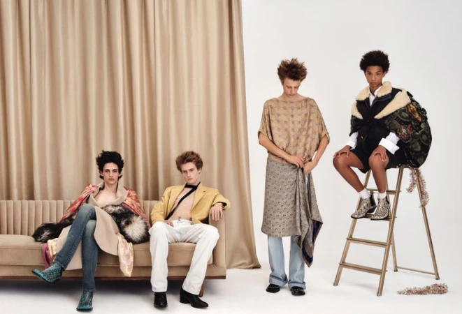 Four male models pose across a tufted sofa and step ladder in eclectic designer menswear against a draped curtain backdrop.