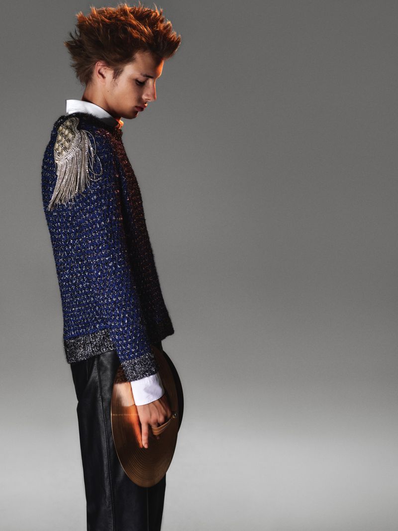 Model in profile wearing a blue textured sweater with silver fringe epaulette and black trousers against a neutral grey studio background.
