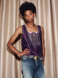 Model posing in a sleeveless purple sequin top and blue denim jeans against draped beige pleated curtains.