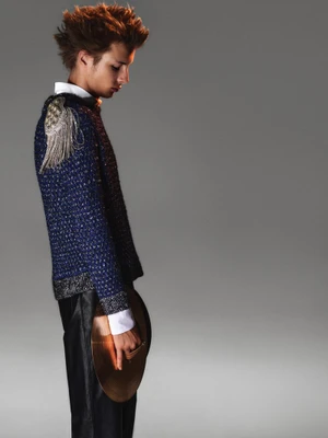 Model in profile wearing a blue textured sweater with silver fringe epaulette and black trousers against a neutral grey studio background.