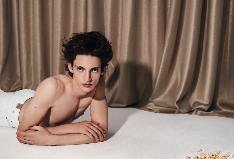 Shirtless young male model with dark hair lying on a white floor against draped beige satin curtains.