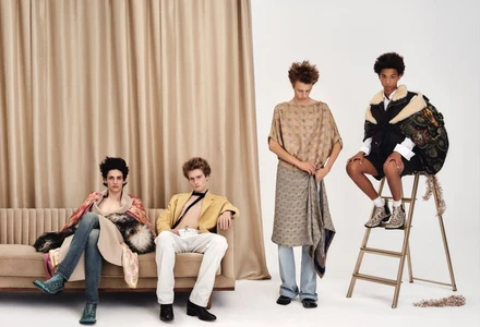 Four male models pose across a tufted sofa and step ladder in eclectic designer menswear against a draped curtain backdrop.